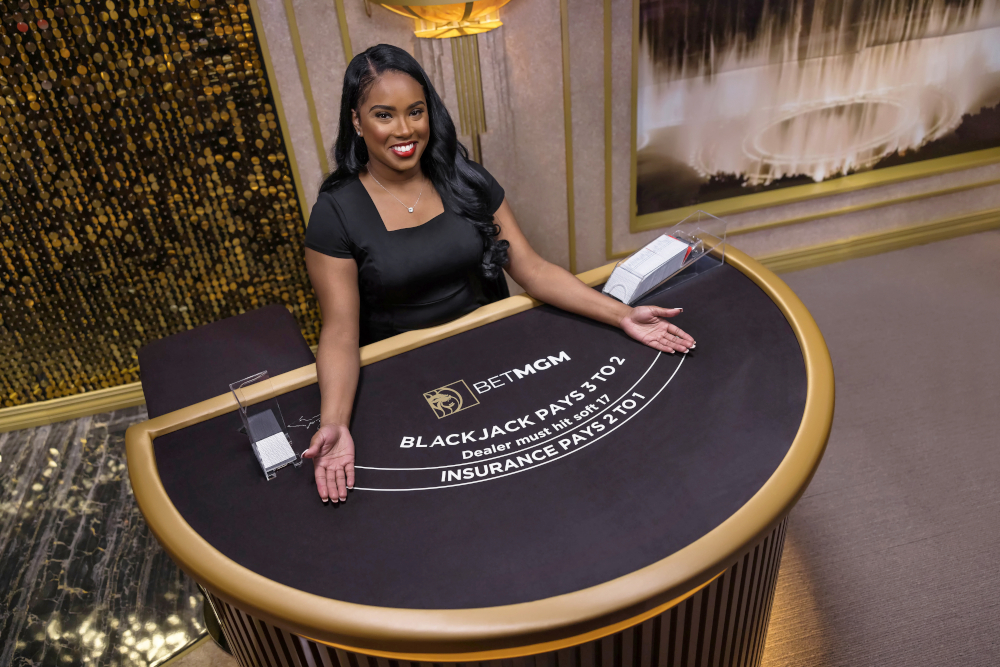 A pretty black woman stands behind a blackjack table with BetMGM brandng on it. Six blackjack tables will broadcast live from new studio in Europe. BetMGM Casino Ontario also adds 108 new slot and video table games to its list of offerings.