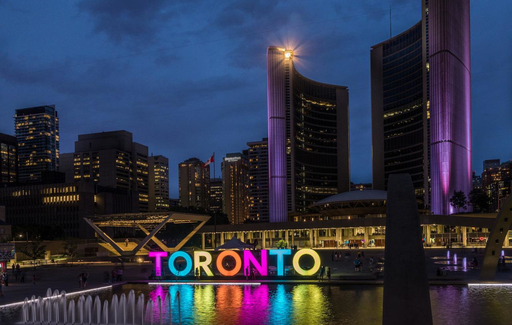 downtown toronto at night. a sculpture spelling out TORONTO is lit up against dark buildings with lights and dark cloudy skies.