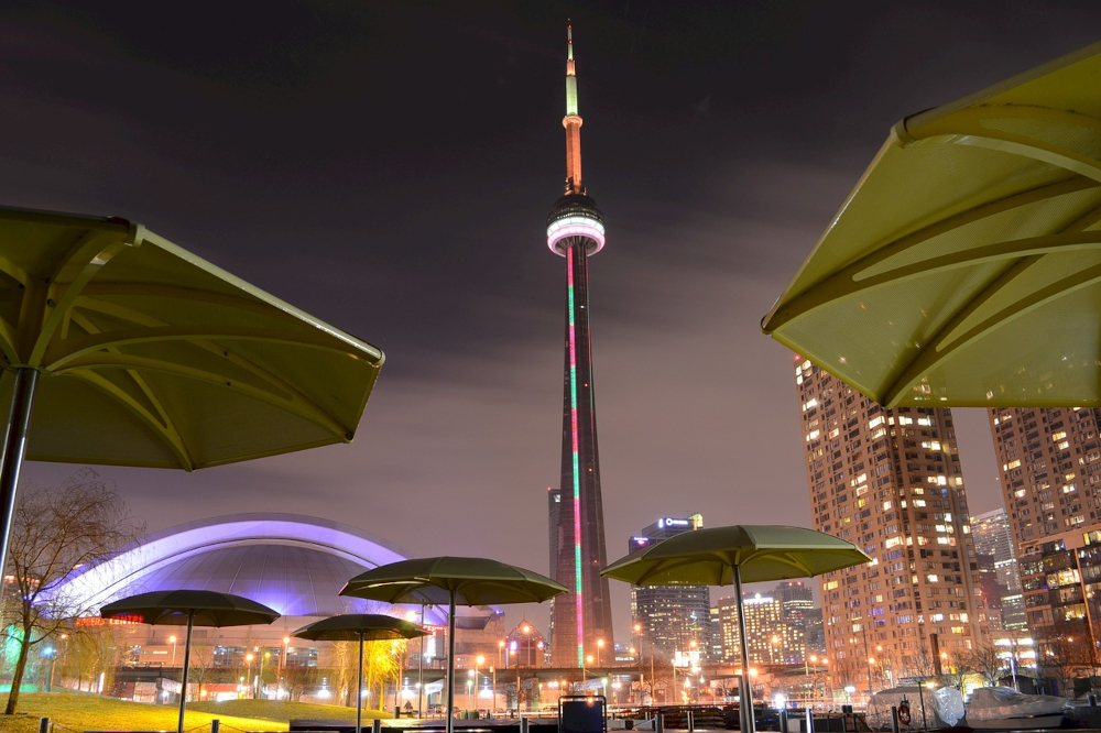A view of downtown Toronto with the CN Tower looming is seen at night, from street level. Lit up buildings and umbrellas are to the sides.