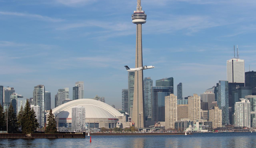 Toronto skyline is seen against blue sky. CN Tower and skyscrapers rise up from the shore of Lake Ontario. in the middle of the image, a plane takes off.